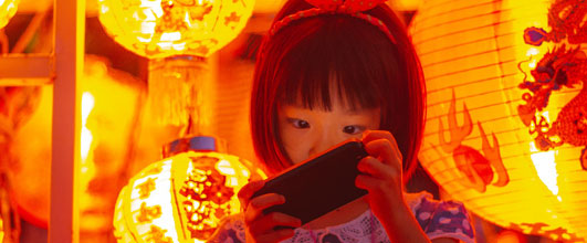 写真素材サイト「PAKUTASO」より、お祭りの提灯とスマオに夢中の女の子。ジユンさんの作品。モデルは、あんじゅさん。 アイキャッチ画像。